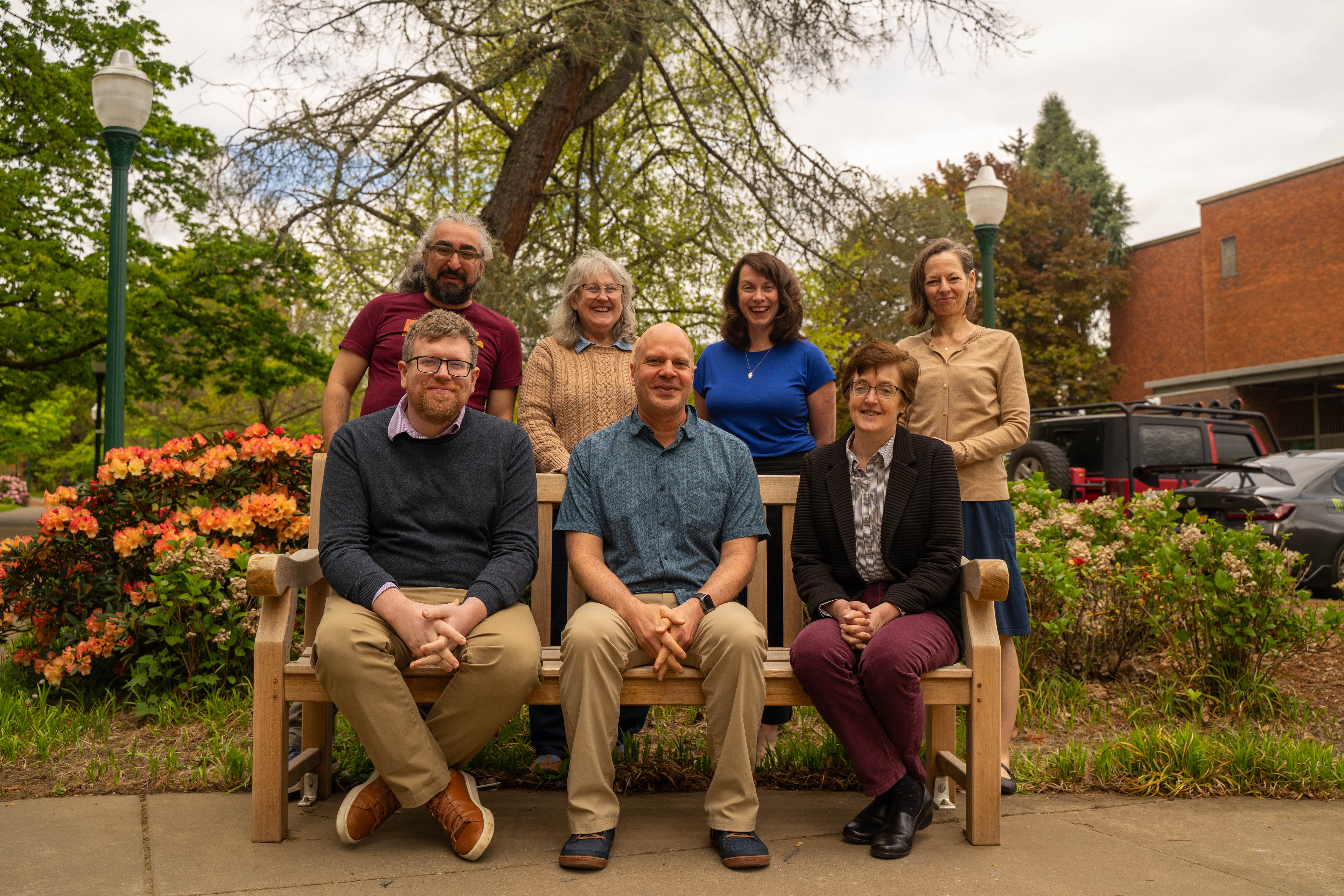 The Teaching Engagement Team, three sitting on an outdoor bench and four standing behind it, on UO's campus