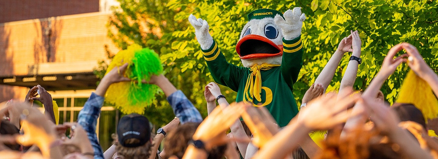 UO duck mascot stands with arms raised in background and in foreground students arms raised