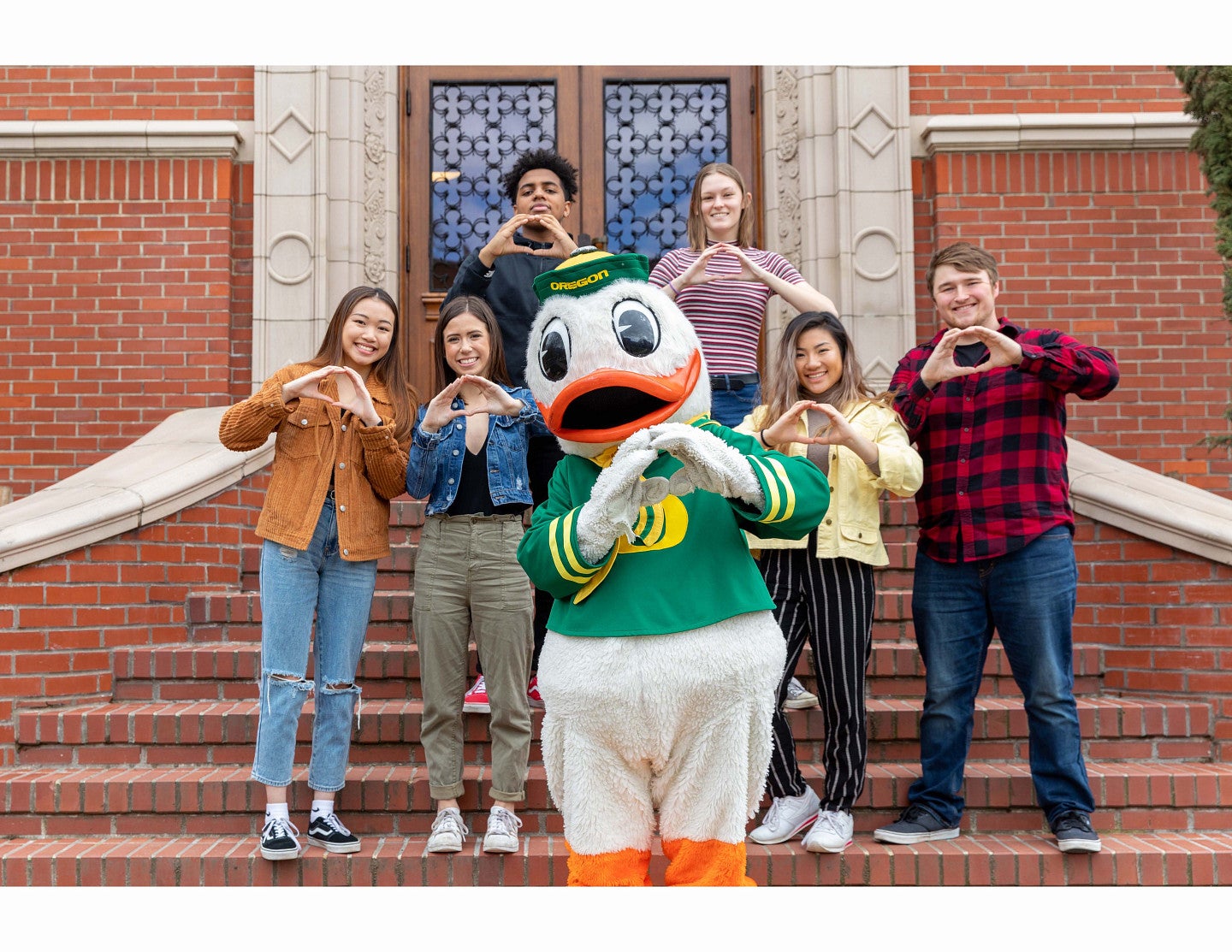 The University of Oregon mascot posing with a diverse group of students on the stairs of a university building.