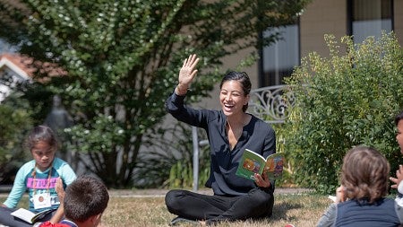Woman seated cross legged on the grass reading to young children