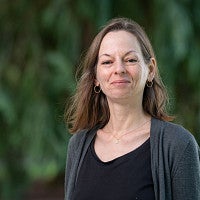 Headshot of white woman with shoulder length brown hair facing camera, smiling, wearing black and grey top and gold earrings, in front of a green background